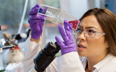 生物处理重组补充剂 身着白大褂、戴着紫色手套的科学家手持并检查一个装有红色液体的透明小瓶。可见实验室设备,包括显微镜的一部分,表明这是一个科学实验室环境