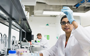 In a modern laboratory with bright white walls and ceilings, a female scientist wearing a white lab coat, blue latex gloves, and black-framed glasses is carefully examining a clear glass test tube filled with a transparent liquid. She holds the test tube up to eye level with a focused expression, ensuring precise observation. The workstation in front of her contains various laboratory glassware, including flasks and beakers filled with solutions, as well as plastic containers with red and yellow lids. Stainless steel faucets and a black lab bench are visible, emphasizing a well-equipped analytical environment. In the background, a male scientist in a white lab coat is working at another station, slightly out of focus, contributing to the dynamic and professional atmosphere of the laboratory. In a modern laboratory with bright white walls and ceilings, a female scientist wearing a white lab coat, blue latex gloves, and black-framed glasses is carefully examining a clear glass test tube filled with a transparent liquid. She holds the test tube up to eye level with a focused expression, ensuring precise observation. The workstation in front of her contains various laboratory glassware, including flasks and beakers filled with solutions, as well as plastic containers with red and yellow lids. Stainless steel faucets and a black lab bench are visible, emphasizing a well-equipped analytical environment. In the background, a male scientist in a white lab coat is working at another station, slightly out of focus, contributing to the dynamic and professional atmosphere of the laboratory.