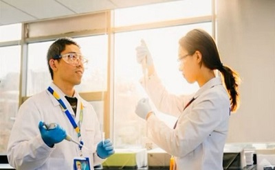 Navigating quality grades Two scientists, a man and a woman, are working in a brightly lit laboratory with large windows allowing sunlight to stream in. The man, wearing safety goggles, a white lab coat, a blue lanyard with an ID badge, and blue nitrile gloves, is holding a pipette and attentively looking at his colleague. The woman, also in a white lab coat and safety glasses, is holding a test tube up to the light with a focused expression, carefully examining its contents. The background features laboratory benches with various equipment, and the golden sunlight casts a warm glow on the scene, emphasizing the collaborative nature of their work.