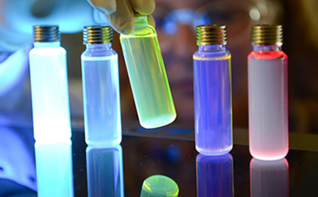 Vials of OLED materials lit up by UV light 在五彩灯光的照耀下,几个带金属盖的玻璃小瓶的特写图像。玻璃瓶中的液体发出蓝色、紫色和红色的光,在反光表面的映衬下,产生了强烈的视觉效果。一只戴着手套的手放在玻璃瓶附近,暗示着科学或实验背景。