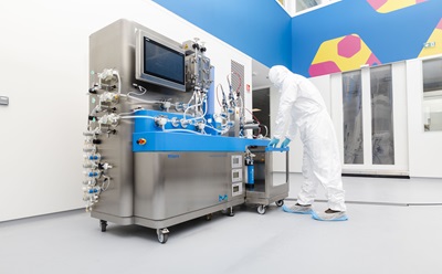 Mobius Multi Column Capture System A technician in a white protective suit and blue shoe covers is operating a large, stainless steel pharmaceutical machine with blue accents, which features various tubes and a digital screen. The background shows a brightly colored, modern laboratory environment with geometric patterns on the walls.