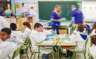 Beyond The Bench A handful of students sit at their desks wearing lab coats and gloves. Two staff volunteers walk around to support students.