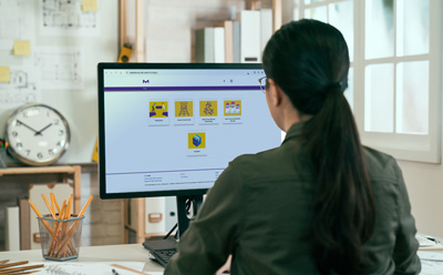 mConfig™ Configurator Home Screen 一位深色头发扎着马尾的女士坐在办公桌前,看着电脑显示器上显示的彩色仪表盘,仪表盘上有六块黄色瓷砖。