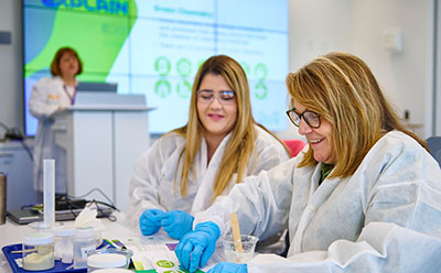 Two scientists at a table doing a demonstration. Members of the Beyond Benign team are engaged in a scientific experiment at a table filled with various lab materials. In the background, a presentation screen displays information related to their work.