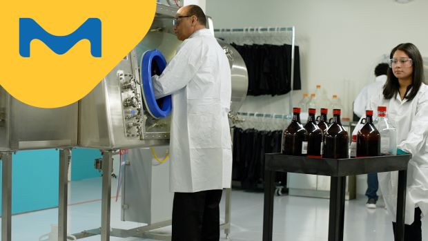 <p>HPAPI成功生产的秘诀</p> A male scientist in a lab coat is using an isolator, while a colleague female scientist in a lab coat is transferring chemicals into the training facility behind him.