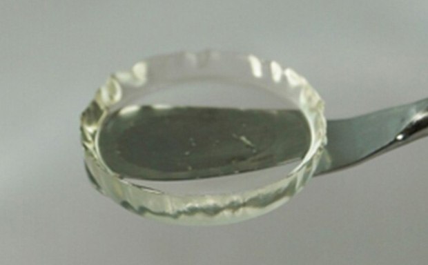 Close-up photo of a transparent, gel-like substance neatly contained within a circular glass dish held by a metal spoon, a sample of a hydrogel 