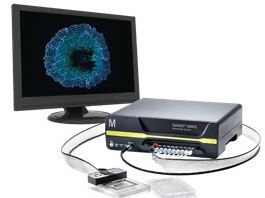 A computer monitor connected to a black electronic device with multiple ports, showcasing a blue circular pattern on the screen. Two small rectangular objects are placed in front of the device, and two cables connect the monitor to the device. 