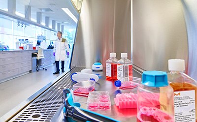 Grey tissue culture hood containing a black handheld cell counter, a square media bottles filled with red fluid, and a cylindrical media bottle filled halfway with red fluid in the foreground. In the middle of the tissue culture hood is a pink tube holder with three clear tubes, a six well clear plate with three wells filled with red media, and two T flasks halfway filled with red fluid. In the background, a white pipette gun, two square media bottles filled with red or clear fluid, and a vortexer. To the left, a male scientist in a white coat walks by.
