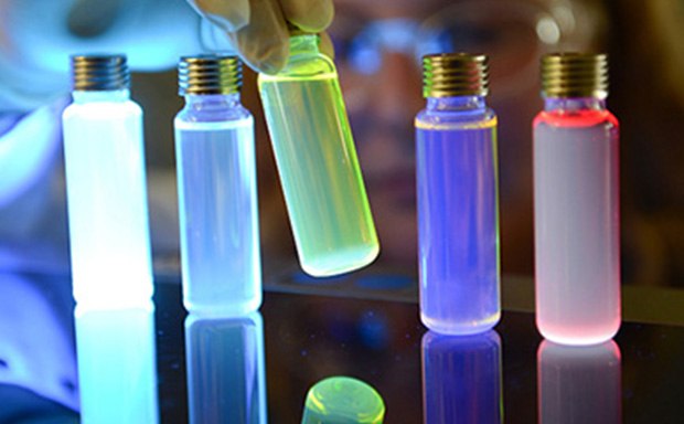 A close-up image of several glass vials with metallic caps, illuminated by colorful lights. The vials contain liquids that glow in shades of blue, purple, and red, creating a visually striking effect against a reflective surface. A gloved hand is positioned near the vials, suggesting a scientific or experimental context.