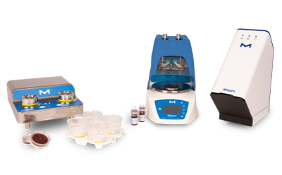 Three pieces of laboratory equipment associated with the Milliflex® Rapid system 2.0. On the left, there’s a stainless steel device with two circular platforms, each holding a clear petri dish with a red medium inside, and several additional petri dishes stacked beside it. In the center, there’s a larger blue device featuring a digital display screen and control buttons; it appears to be an incubator or analyzer with an open door revealing internal components. On the right side of the image stands a white device.