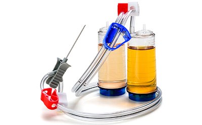 A Steritest NEO Membrane Filtration System. This system consists of a filtration unit with two connected chambers filled with a yellowish liquid. The left chamber has a red clamp, and the right chamber has a blue clamp; both are mounted on stands. A needle attached to additional tubing is also present.