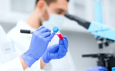 A person in a white lab coat stands in a laboratory facing away from the camera, holding a clipboard and writing notes. The laboratory is equipped with shelves stocked with various lab equipment such as beakers, flasks, test tubes, and bottles containing chemicals labeled with different colors.