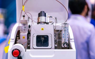 A detailed view of a protein mass spectrometry machine, featuring various components such as tubes, wires, and dials, with a visible emergency stop button and cautionary yellow label. In the background, a scientist in a lab coat works within a laboratory setting, highlighting the machine’s use in scientific research and technological advancement.