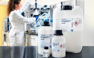 A scientist in a lab coat working with equipment and chemicals in a laboratory setting. In the foreground, there are three chemical containers with labels, two are large and one is smaller, marked with hazard symbols for flammable and corrosive substances.