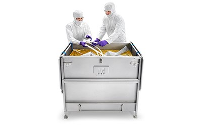 Two individuals in protective white suits, gloves, and head covers are standing at a stainless steel cart with large gold-colored bags inside it.
