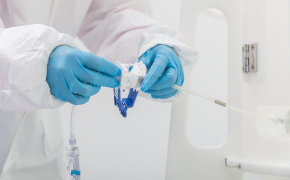A person in protective gear, including gloves, handling a medical syringe and vial. The individual is dressed in white, with blue gloves, and is focused on the medical equipment.