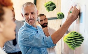 A group of people, one of whom is writing on a whiteboard, while others observe, with colorful geometric shapes displayed in the background.