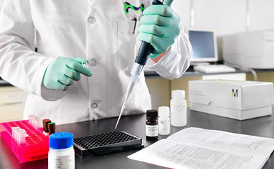 Bioactive Small Molecule Inhibitors A laboratory technician in a white lab coat and green gloves is using a pipette to transfer liquid into a black microplate. Various laboratory supplies, including bottles of reagents and a pink rack for test tubes, are visible on the table, alongside a document with printed instructions. In the background, laboratory equipment is partially visible.