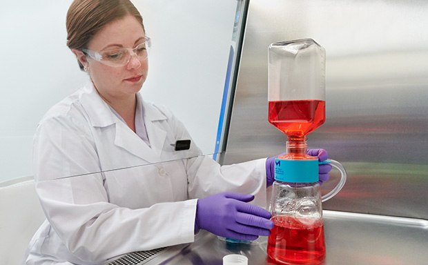  A woman in a white lab coat, working with laboratory flask containing a red liquid, which is connected to another piece of equipment with blue liquid at the bottom.