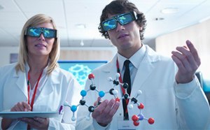 Two scientists, a woman and a man, stand side by side in a brightly lit, modern laboratory wearing white lab coats and large black 3D glasses. The woman, with straight blonde hair, holds a tablet in both hands and wears a red lanyard. The man has short brown curly hair and is holding a colorful molecular model made of plastic balls and sticks in black, blue, white, red, and purple, representing different atoms and bonds. He also wears a red lanyard and a name badge over a white shirt and black tie. Behind them, a digital screen displays a bright blue hexagonal molecular structure. The room is well-lit with soft white ceiling lights, pale wood walls, and a partially visible computer station in the background, giving the setting a clean and high-tech atmosphere focused on molecular research or visualization. 