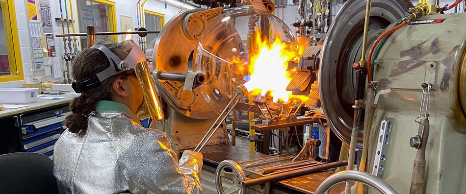 Glassblower works on flask A round-bottom flask sits in a lathe and a glassblower directs a targeted flame onto the flask’s neck. The glassblower wears protective clothing, including a specialized jacket, gloves and face shield.