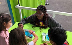 Curiosity Cube A group of children is engaged in a hands-on science activity at a bright green table, where they are exploring a water filtration experiment. The facilitator, wearing a SPARK™ shirt, demonstrates the process while the children attentively observe and participate, fostering an interactive learning experience outdoors.