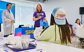 Curiosity Labs™ A child wearing a "Thinking Cap" hat is seated at a table, facing a presentation about water filtration, with two adults standing in front of a large screen displaying relevant information. The adults are engaged in explaining the topic, while various educational materials, including cups and worksheets, are visible on the table, creating an interactive learning environment.