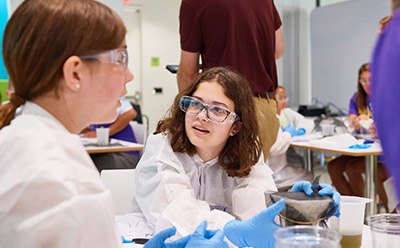 Curiosity Programming Two children are engaged in a hands-on science activity, wearing protective lab coats and gloves. One child, holding a container, is demonstrating something to another child who is attentively listening, while a few adults and other participants are visible in the background, creating an interactive and educational environment.