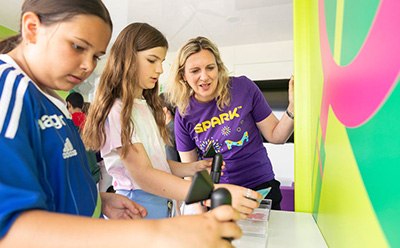 SPARK™ Three children are engaged in an interactive activity at a colorful educational booth, with one child holding a device while another observes intently. An adult, wearing a purple SPARK™ volunteer shirt, is guiding them through the activity, promoting hands-on learning and engagement. The vibrant background features bright colors and patterns, enhancing the lively atmosphere of the event.