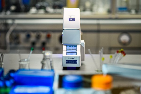 A close-up view of the Penn PhD Photoreactor M2, a laboratory instrument with a sleek white design and a digital display, positioned on a lab countertop surrounded by various laboratory glassware and equipment, including colorful reagent bottles and pipettes in the background.